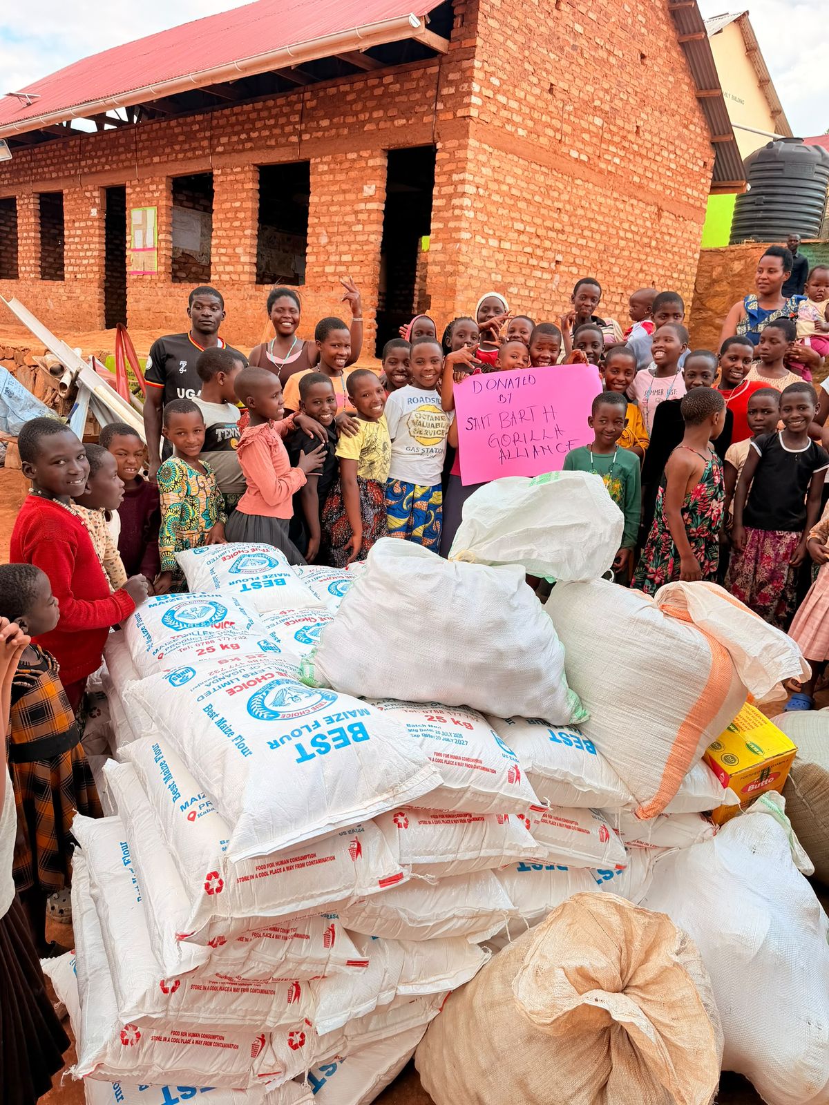 Children and community members gather around a large delivery of maize flour and food sacks outside a brick school building, holding a pink sign