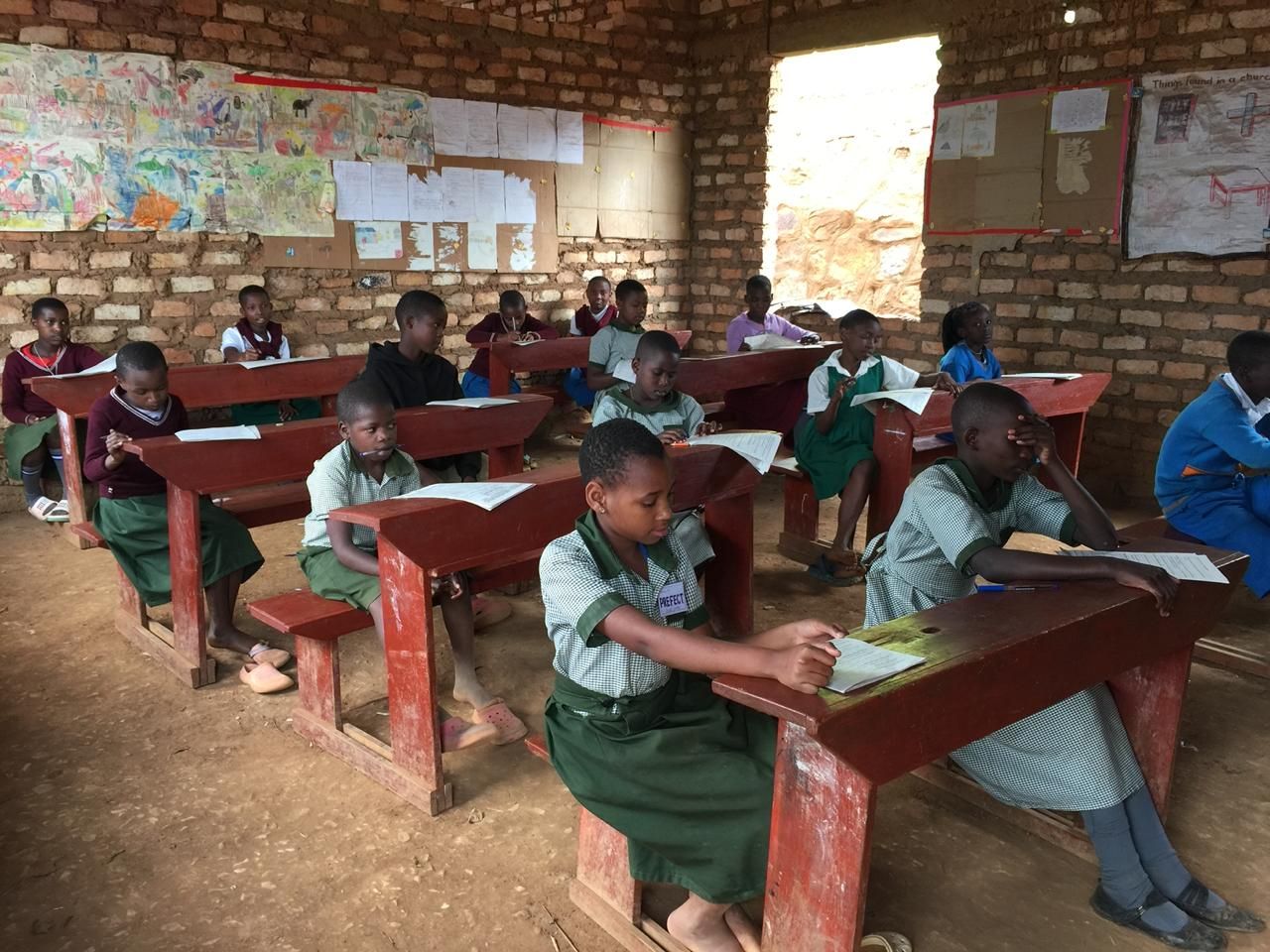 Pupils in green uniforms sit at red wooden desks writing in a brick classroom